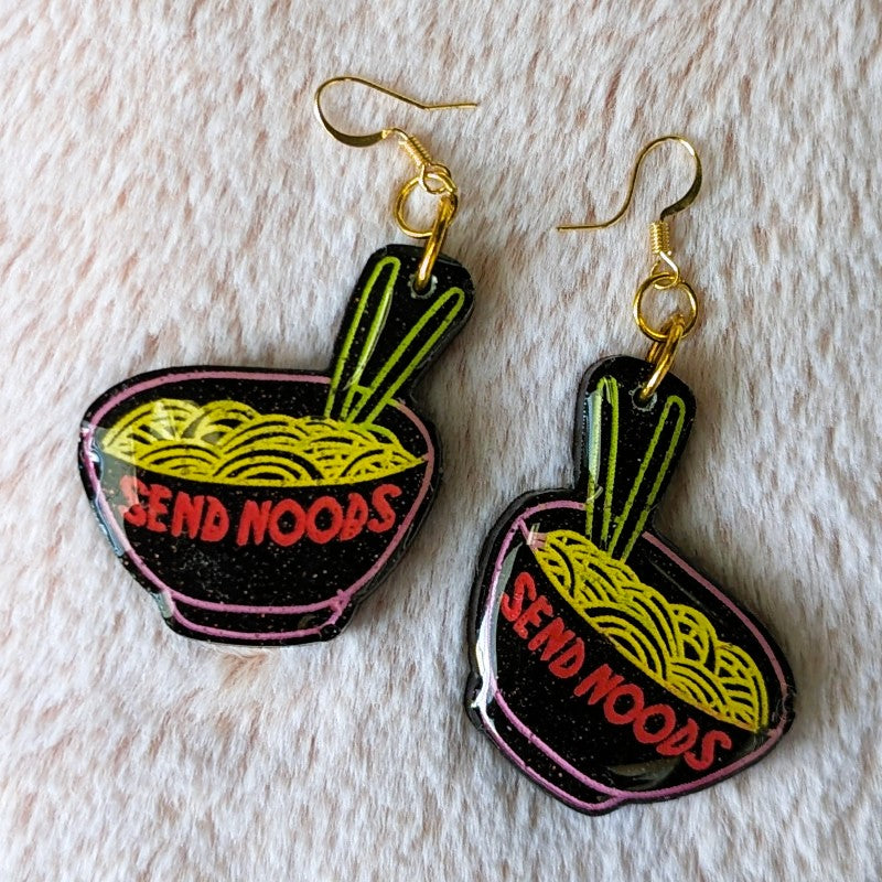 funny and glittery earrings of ramen bowls that say "send noods"
