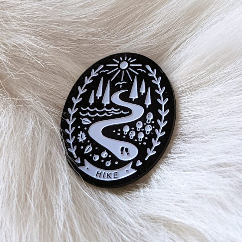 enamel pin of a walking path through a forest scene that says "hike"