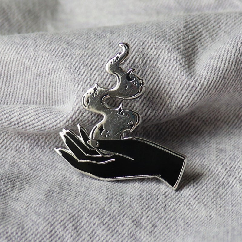 enamel pin featuring a hand with mystical smoke coming out of it