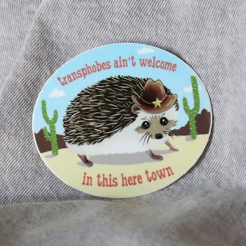 cute sticker of a hedgehog in an old west costume and the words "transphobes ain't welcome in this here town"