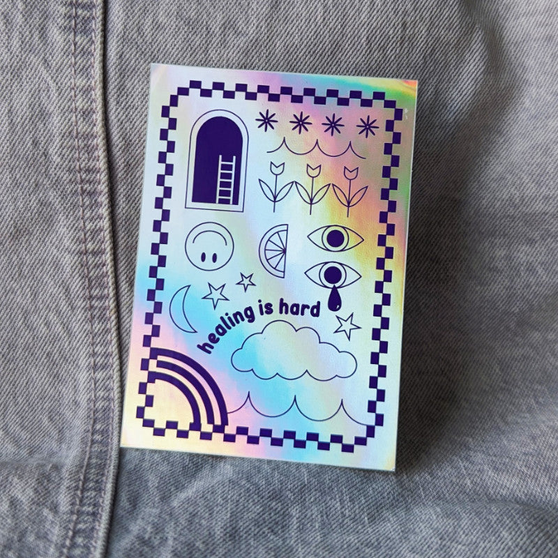 cool holographic sticker that says "healing is hard" with a funky illustration of clouds, eyes, flowers, and more