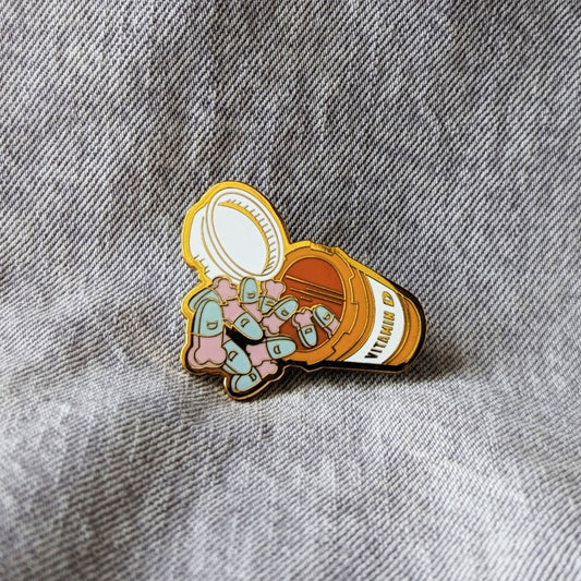 funny enamel pin of a pill bottle that says "vitamin d" and penis-shaped pills spilling out of it