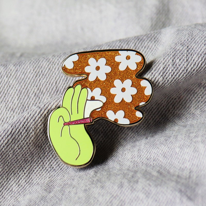 enamel pin of a hand, a joint, and a plume of glittery smoke with a flower design