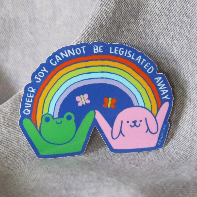 sticker of a frog, puppy, and rainbow and the words "queer joy cannot be legislated away"