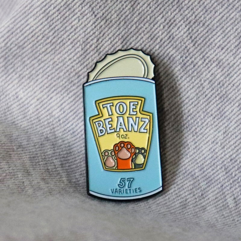 enamel pin depicting an open can with kitty paws and the words "toe beans"